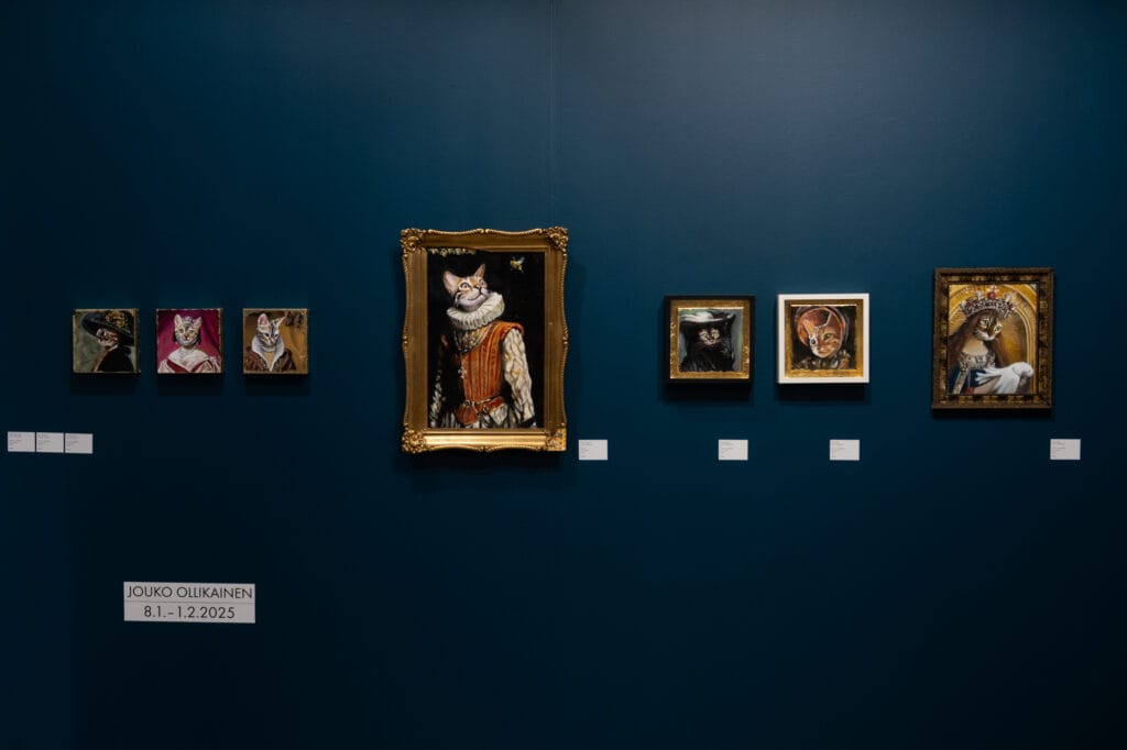 Maalauksia galleriassa, kissakasvot ja kehyksissä.