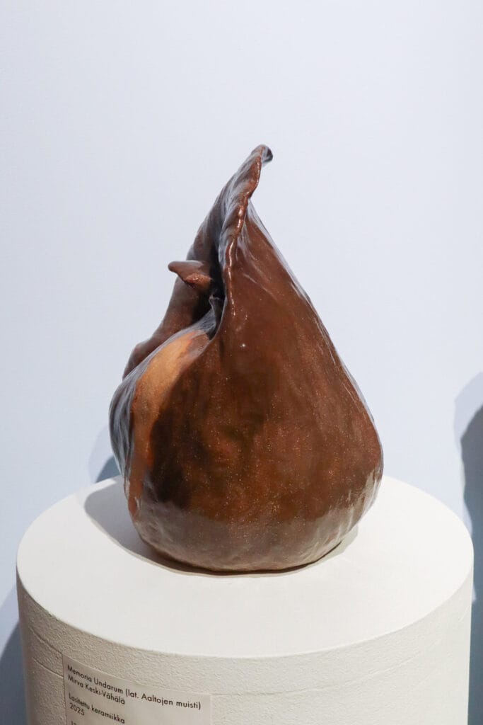 Ruskea keraaminen veistos näyttelypöydällä valkoisella taustalla.